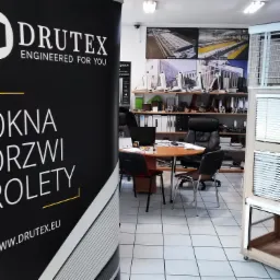 Wnętrze biura firmy z ekspozycją produktów: okien, drzwi i rolet, widoczny baner reklamowy z logo firmy i hasłem 'Engineered for you'.