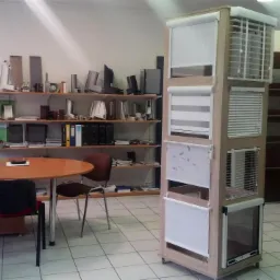 Ekspozycja różnych typów osłon okiennych, w tym żaluzji i rolet, prezentowana na drewnianym stojaku w biurze z widocznym biurkiem, krzesłami i półkami w tle.