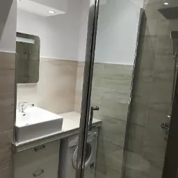 Nowoczesna łazienka z kabiną prysznicową, umywalką nablatową na szafce z szufladami i pralką w zabudowie, wykończona płytkami w odcieniach beżu i szarości.