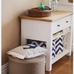 Biała konsola z drewnianym blatem, wiklinowym koszem, szklanymi butelkami, pufą i poduszką z motywem kotwicy w aranżacji wnętrza w stylu marynistycznym.