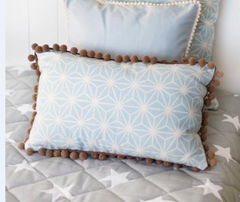 Dwie poduszki dekoracyjne: prostokątna z geometrycznym wzorem i brązowymi pomponami na krawędzi oraz kwadratowa z białymi pomponami, ułożone na szarej narzucie w białe gwiazdy.