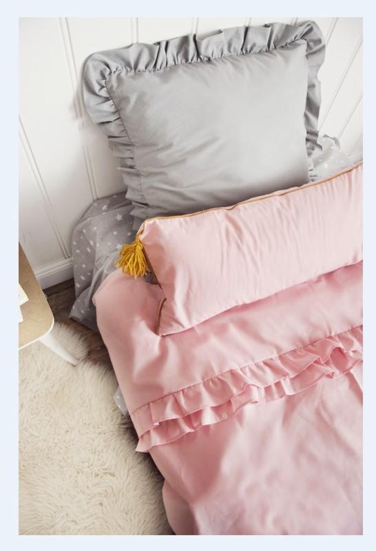 Łóżko dziecięce z szarą poduszką z falbanką, różową poduszką z pomponem i różową pościelą z falbanami, na tle białej boazerii i puszystego dywanu.