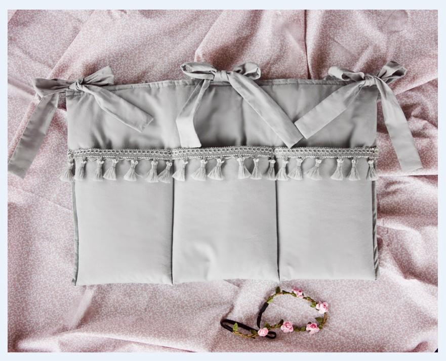 Szary, tekstylny organizer do łóżeczka dziecięcego z trzema kieszeniami, ozdobiony frędzlami i wiązaniami, leżący na różowej pościeli w drobny wzór, obok opaska z różowymi kwiatkami.