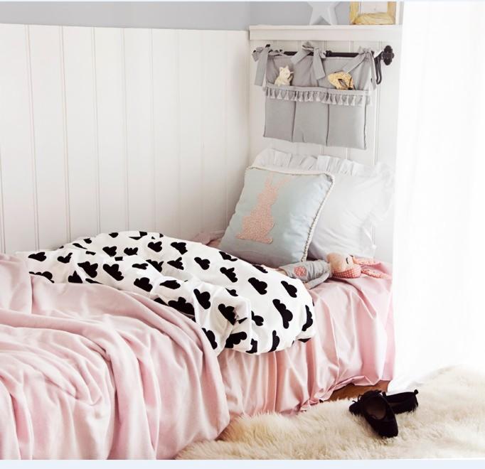Dziecięce łóżko z białym zagłówkiem z pionowych paneli, różową pościelą, czarno-białą kołdrą w chmurki i szarą wiszącą kieszenią na drobiazgi z zawartością.