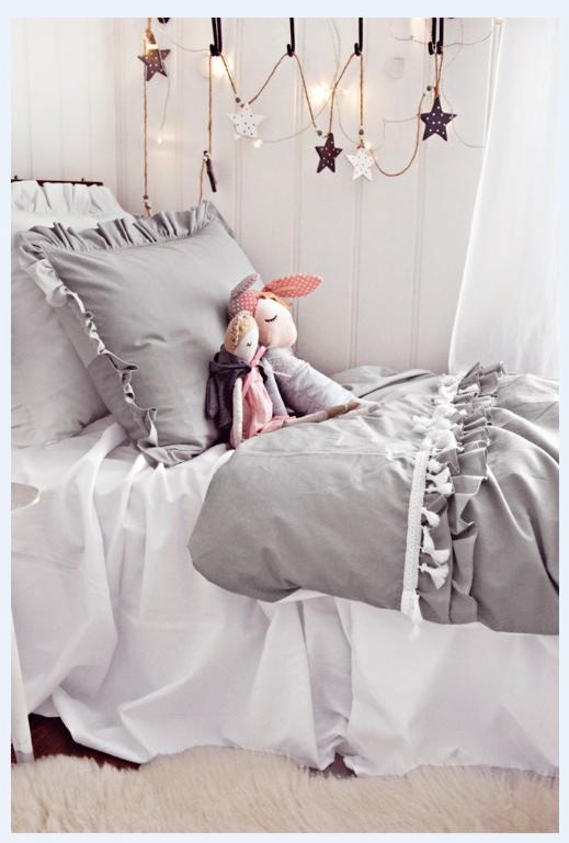 Łóżko dziecięce z szarą pościelą z falbanami i dekoracją w postaci gwiazdek, na którym siedzą dwie lalki.
