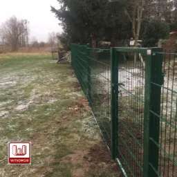 Zielona brama w systemie ogrodzeniowym panelowym na działce z resztkami śniegu, widok z boku na ogrodzenie.