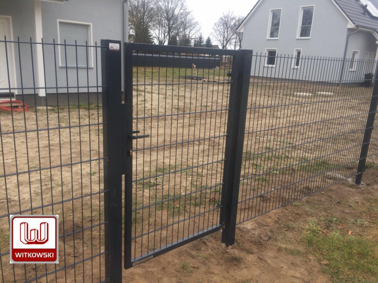 Ciemnoszara, otwarta furtka w systemie ogrodzenia panelowego z widocznym logo firmy Witkowski w lewym dolnym rogu, na tle szarego domu.