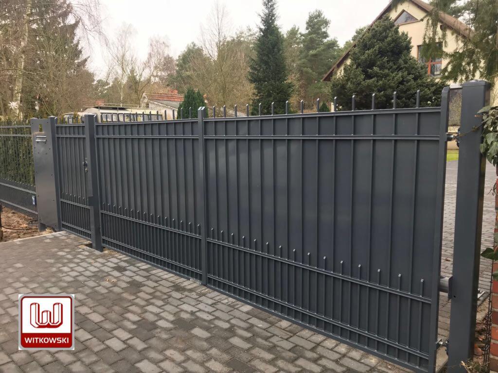 Szara, metalowa brama wjazdowa z pionowymi prętami, widoczna z bliska na tle brukowanej nawierzchni i domu.