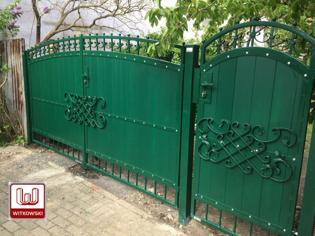 Zielona brama wjazdowa z ozdobnymi elementami kutymi, widoczny fragment chodnika i roślinność w tle.