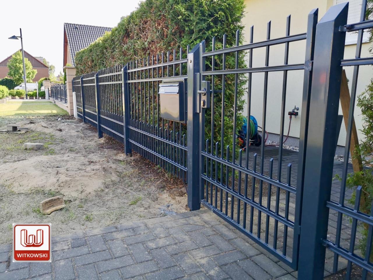 Ciemnoszara brama wjazdowa z metalowymi prętami i skrzynką na listy, widoczna z zewnątrz posesji na tle zielonego żywopłotu.