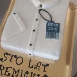 Tort w kształcie białej koszuli z mankietem i kołnierzykiem, z czarnymi guzikami i etykietą 'REMI 2019', leżący na złotej tacy z napisem 'STO LAT REMIGIUSZ' wykonanym czarnym lukrem.