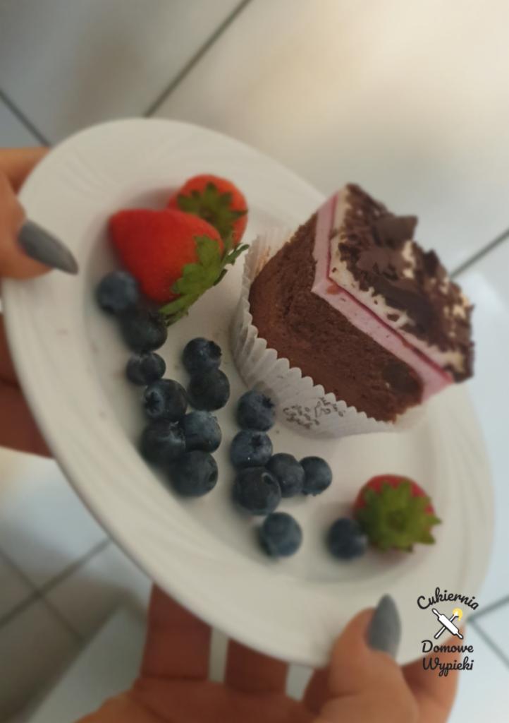 Kawałek tortu czekoladowego z kremem różowym, ozdobiony borówkami i truskawkami na białym talerzu, trzymanym w dłoniach z szarym manicure.