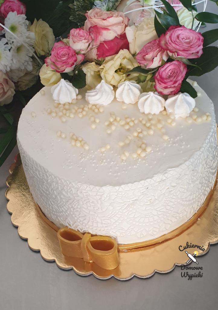 Tort śmietankowy z dekoracją z białych bezików, perełek i żywych róż, na złotej podkładce z dekoracyjną kokardą.