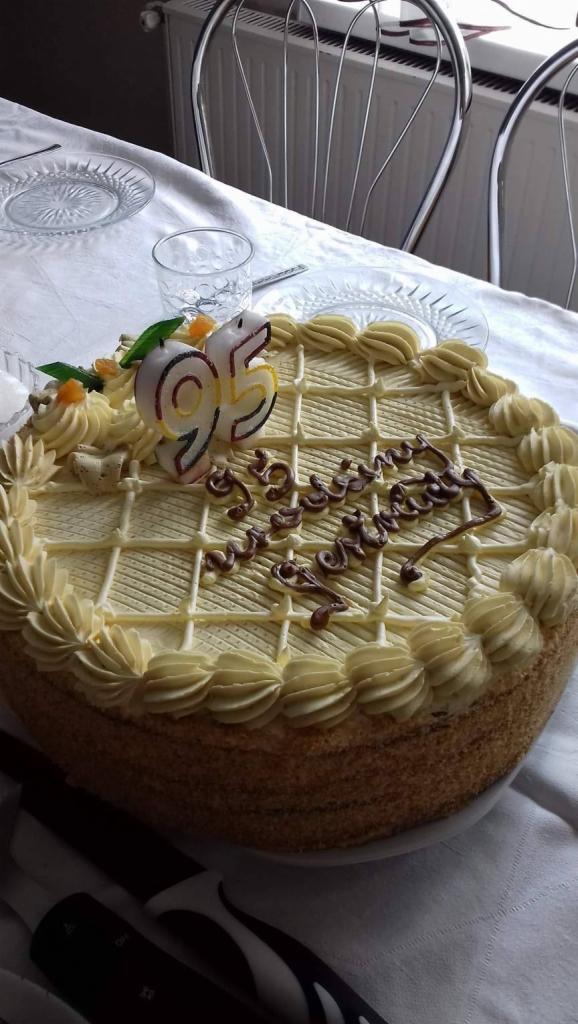 Tort urodzinowy z kremem, dekoracją z czekolady i świeczką w kształcie liczby 95, ustawiony na stole z nakryciem obok szklanki i talerza.