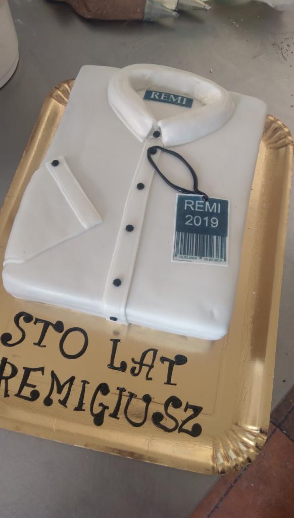 Tort w kształcie białej koszuli z mankietem i kołnierzykiem, z czarnymi guzikami i etykietą 'REMI 2019', leżący na złotej tacy z napisem 'STO LAT REMIGIUSZ' wykonanym czarnym lukrem.