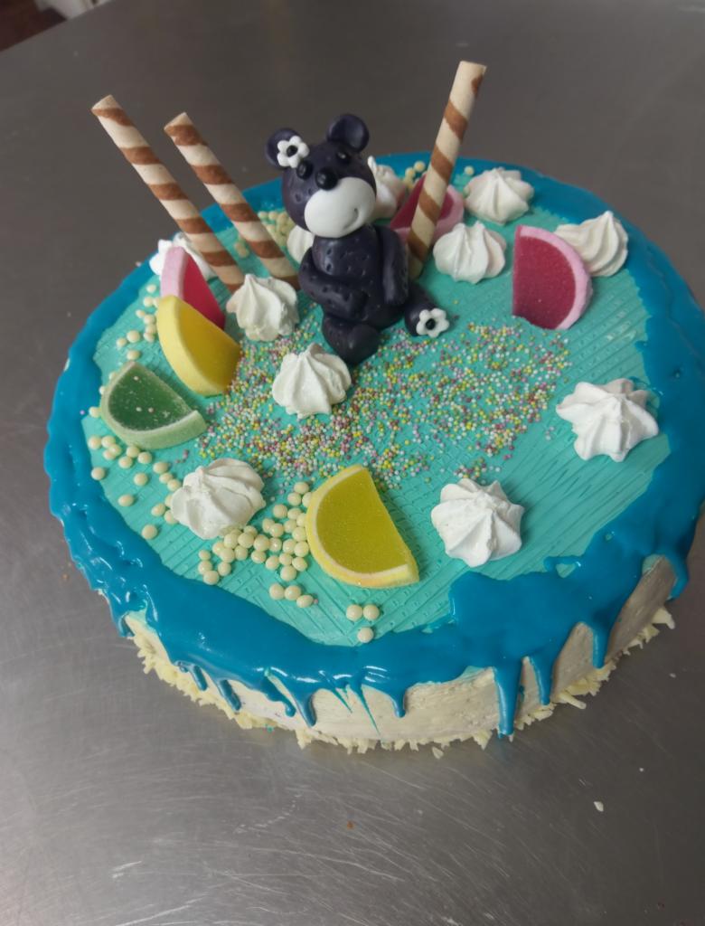 Tort urodzinowy z turkusowym lukrem, dekorowany cukierniczym misiem, bezami, waflami i żelkami w kształcie owoców.