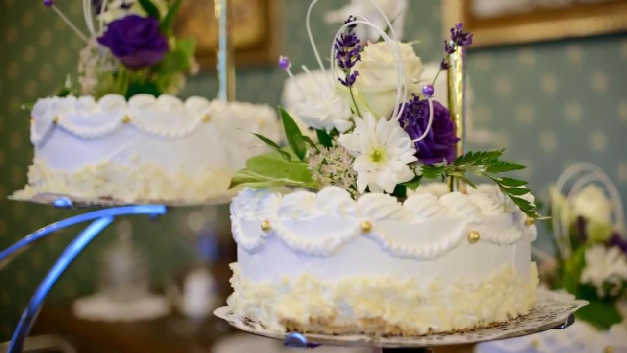 Dwa torty na paterach, udekorowane białym kremem, złotymi kuleczkami i kompozycjami z białych i fioletowych kwiatów oraz zielonych liści, z widoczną fakturą tła.