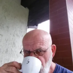 Mężczyzna z siwą brodą pije kawę z białego kubka z czarnym symbolem, na tle ceglanej kolumny i jasnej ściany.