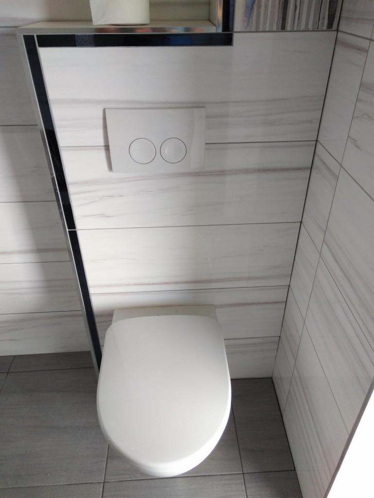 Nowoczesna toaleta wisząca z białym przyciskiem spłukującym na ścianie wyłożonej płytkami imitującymi marmur, szara podłoga z płytek, czarna listwa dekoracyjna.