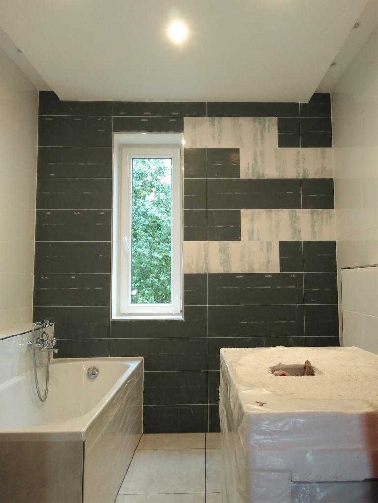 Łazienka w trakcie prac wykończeniowych; widoczna wanna, okno i częściowo wyłożone płytkami ściany z elementami dekoracyjnymi oraz zabudowa w folii ochronnej.