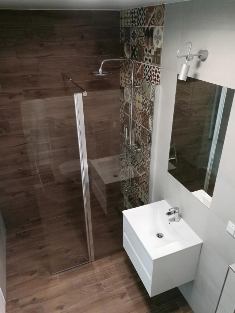 Nowoczesna łazienka z kabiną prysznicową typu walk-in, brązowymi płytkami imitującymi drewno, dekoracyjną ścianą z patchworkowych płytek oraz białą umywalką nablatową z szafką.