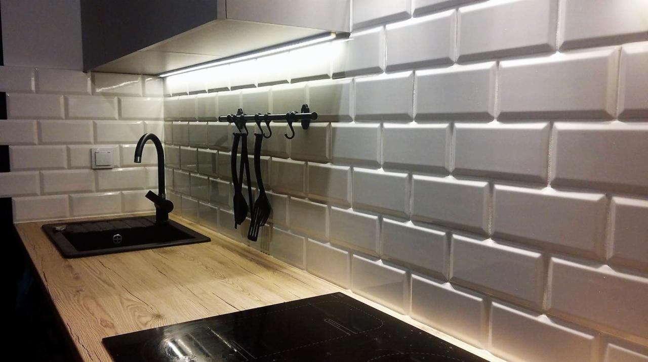 Białe, błyszczące płytki typu metro, czarna bateria i zlew, drewniany blat podświetlony listwą LED w nowoczesnej kuchni.