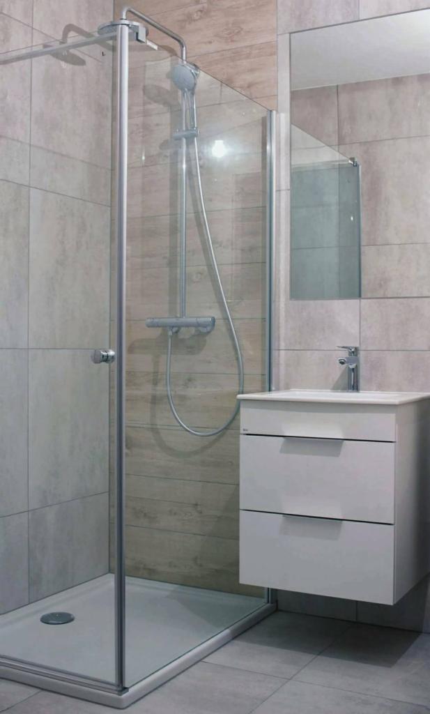 Nowoczesna łazienka z kabiną prysznicową typu walk-in, umywalką z szafką w kolorze białym i dużym lustrem, wykończona płytkami w odcieniach szarości i beżu imitującymi drewno.