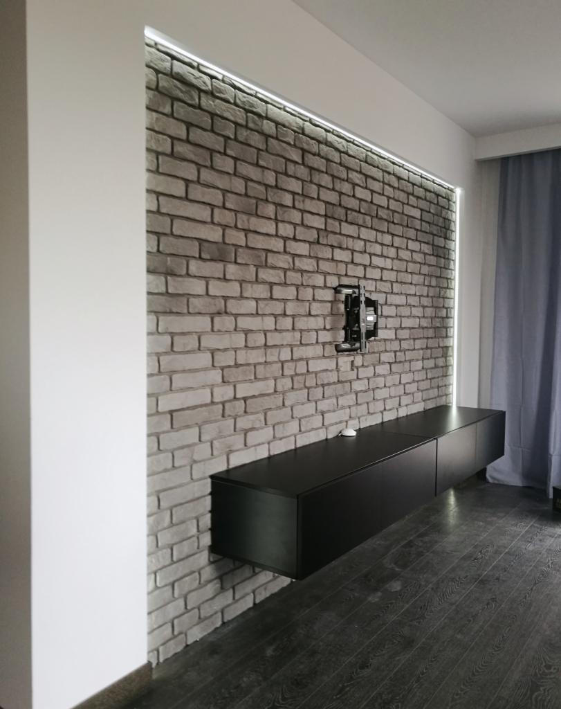 Nowoczesne wnętrze: ściana z szarej cegły z podświetleniem LED, wisząca czarna szafka RTV i drewniana podłoga w ciemnym odcieniu, uchwyt do telewizora na ścianie.
