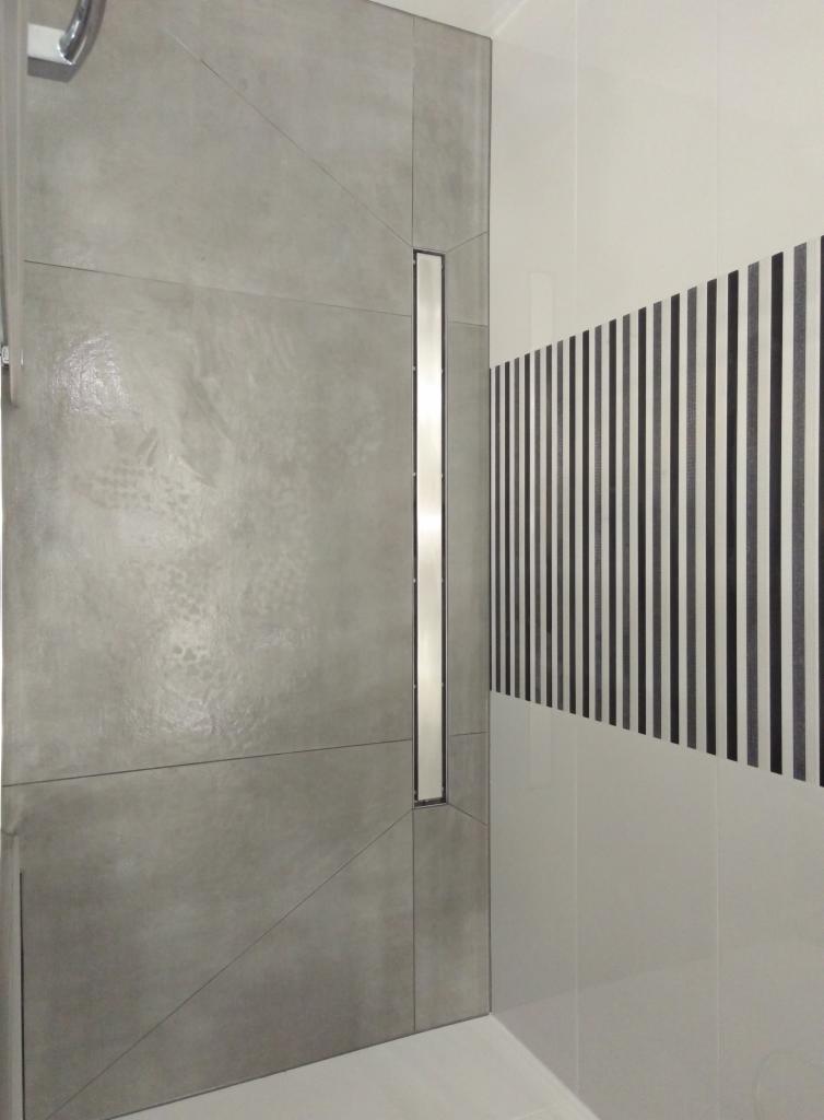 Nowoczesna łazienka z szarą ścianą wykończoną betonem architektonicznym, liniowy odpływ prysznicowy i białe płytki z czarnymi pionowymi pasami dekoracyjnymi.