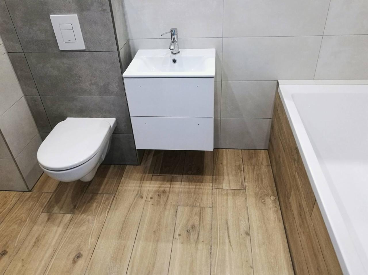 Nowoczesna łazienka z podwieszaną toaletą, szafką z umywalką i wanną, wykończona płytkami imitującymi drewno na podłodze i szarymi płytkami na ścianach.