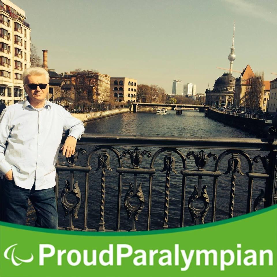 Mężczyzna w okularach przeciwsłonecznych opiera się o metalową balustradę mostu z widokiem na rzekę i architekturę Berlina, w tle wieża telewizyjna; na dole zielony baner z napisem ProudParalympian.