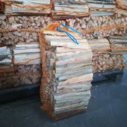 Ułożone pakiety pociętego drewna opałowego, zabezpieczone pomarańczową siatką i niebieskim sznurkiem, na szarym betonowym podłożu.