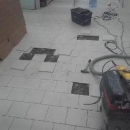 Demontaż uszkodzonych płytek podłogowych, widoczny młotek, szlifierka kątowa i odkurzacz przemysłowy.