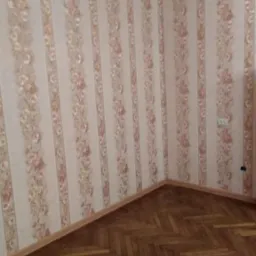 Pusty pokój z tapetą w kremowe pasy z motywem róż i parkietem ułożonym w jodełkę, widoczny fragment listwy przypodłogowej i kontakt.