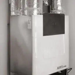 Biała jednostka centralna wentylacji mechanicznej z odzyskiem ciepła, podłączona do przewodów wentylacyjnych w izolacji aluminiowej, umieszczona na metalowej podstawie w rogu pomieszczenia.