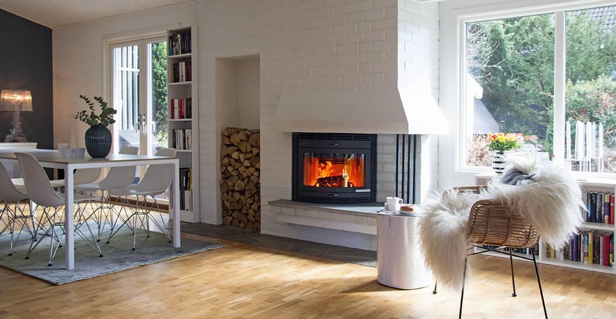 Salon z paleniskiem kominkowym w obudowie z białej cegły, drewnem obok i płomieniami wewnątrz, w jasnym wnętrzu z drewnianą podłogą, stołem i krzesłami.