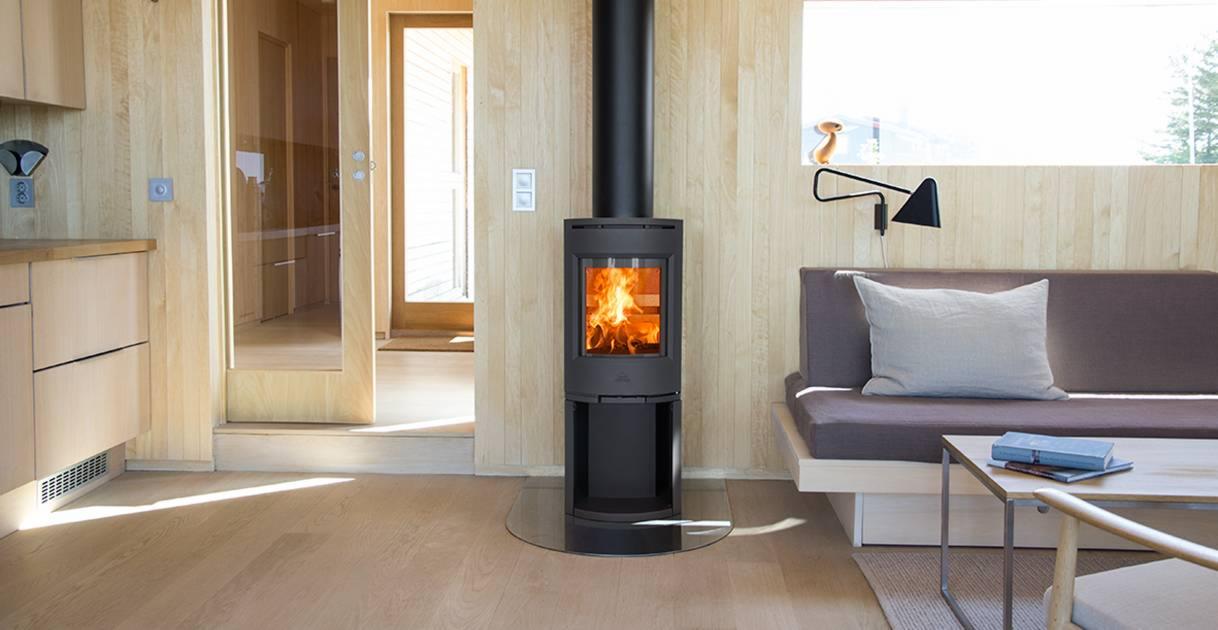 Nowoczesny, czarny piec wolnostojący z paleniskiem, umieszczony w minimalistycznym wnętrzu z drewnianymi panelami, szklaną podstawą i widocznym płomieniem.