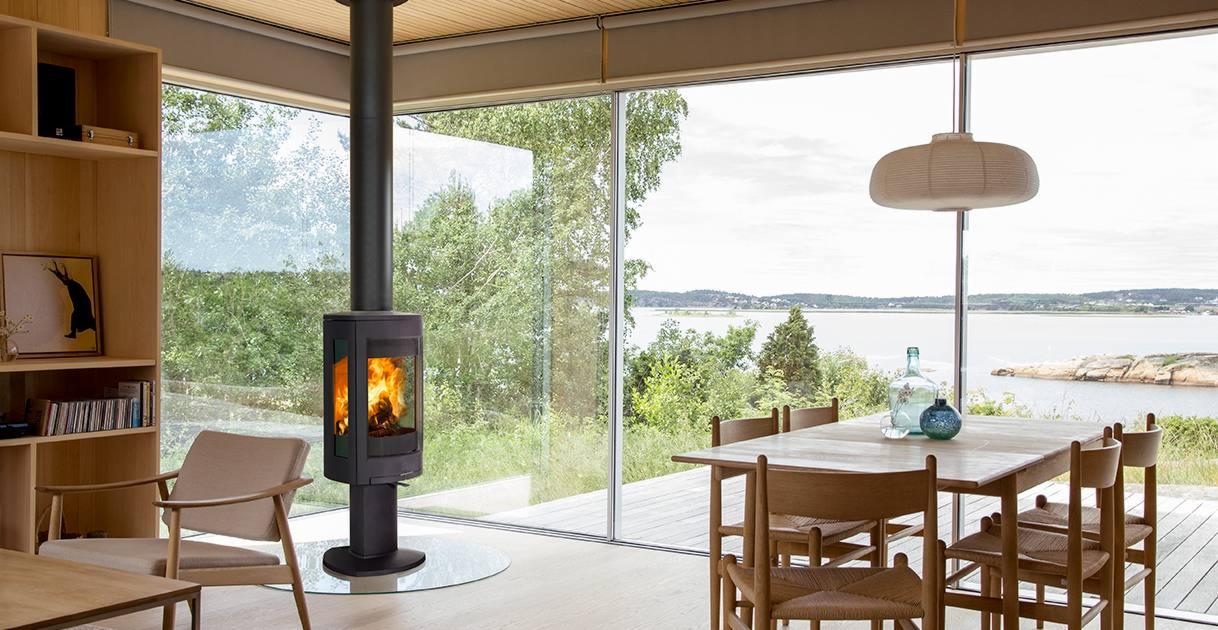Nowoczesny, czarny kominek wolnostojący z paleniskiem, umieszczony na szklanej podstawie w przestronnym salonie z panoramicznym oknem z widokiem na jezioro.