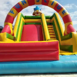 Kolorowy dmuchany zamek ze zjeżdżalnią i motywem klauna na tle błękitnego nieba z chmurami, widoczny fragment budynku z panelami słonecznymi.