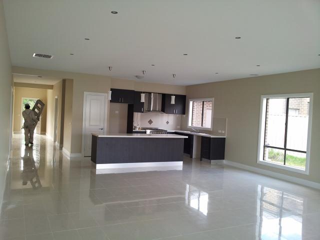 Przestronne, jasne wnętrze domu w trakcie wykańczania, z błyszczącą podłogą, nowoczesną kuchnią z wyspą i pracownikiem w oddali niosącym składaną drabinę.