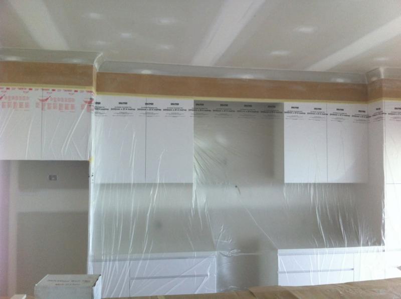 Białe szafki kuchenne zabezpieczone folią ochronną podczas prac wykończeniowych; widoczne taśmy malarskie i listwy sufitowe.