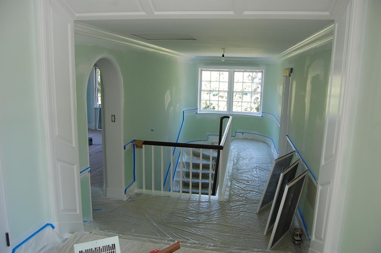 Wnętrze domu w trakcie malowania na jasnozielono, zabezpieczone folią i taśmą malarską, widoczne schody i okno.