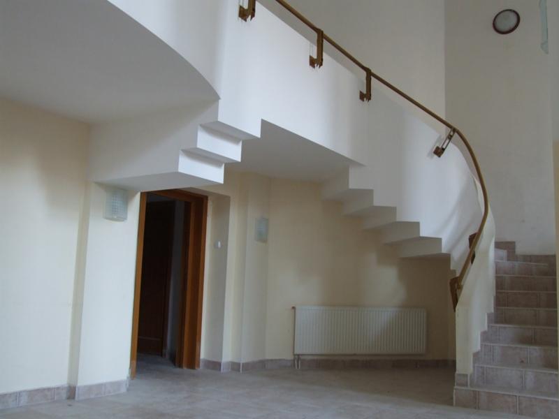 Klatka schodowa w trakcie prac wykończeniowych: widoczne schody z płytek, surowe ściany w odcieniach beżu, drewniane drzwi oraz grzejnik.