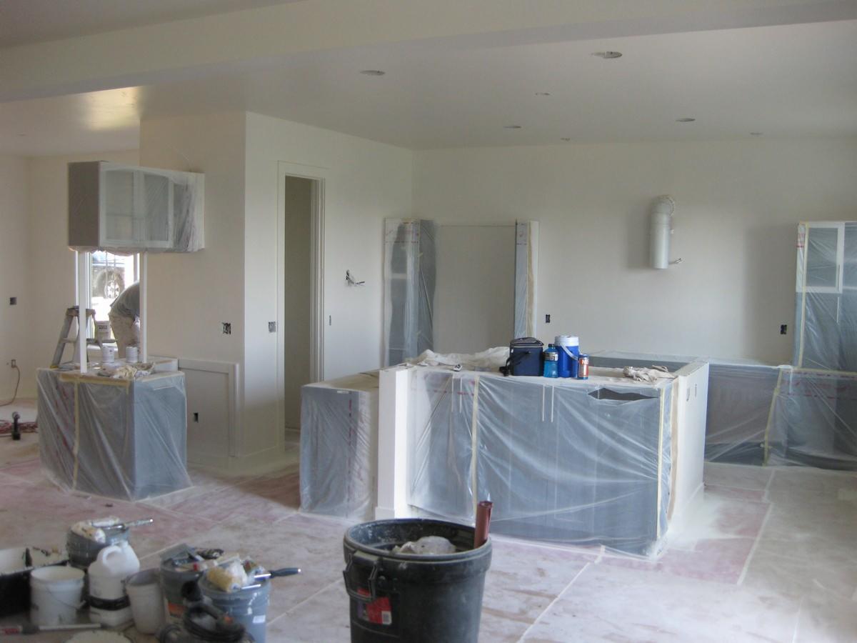 Wnętrze kuchni w trakcie malowania: meble i podłoga zabezpieczone folią, widoczne pędzle, wiaderka z farbą, drabina i pracownik w tle.