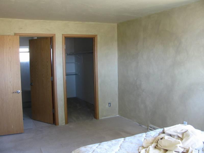 Pokój przed odświeżeniem: widoczne dwie drewniane ościeżnice z drzwiami, jedna otwarta, druga do garderoby z półkami, ściana z dekoracyjnym tynkiem w odcieniach beżu.