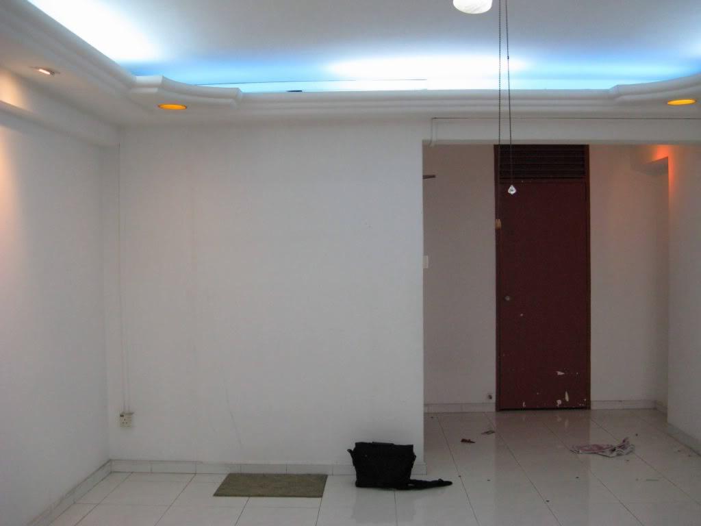 Puste pomieszczenie z białymi ścianami, białymi kafelkami na podłodze i podwieszanym sufitem z oświetleniem LED, widoczny brązowy futryna drzwi i czarny worek na podłodze.