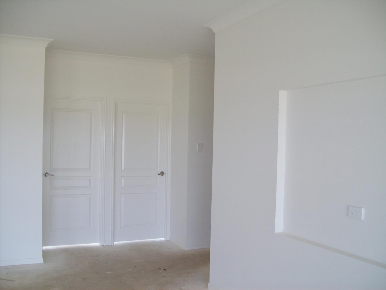 Jasne wnętrze w trakcie wykańczania, białe ściany, dwoje białych drzwi z panelami, widoczne gniazdko elektryczne i wnęka w ścianie, surowa podłoga.