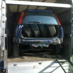 Niebieski samochód osobowy z czterema oponami w bagażniku, zabezpieczony pasami wewnątrz skrzyni ładunkowej samochodu dostawczego z otwartą rampą załadunkową.