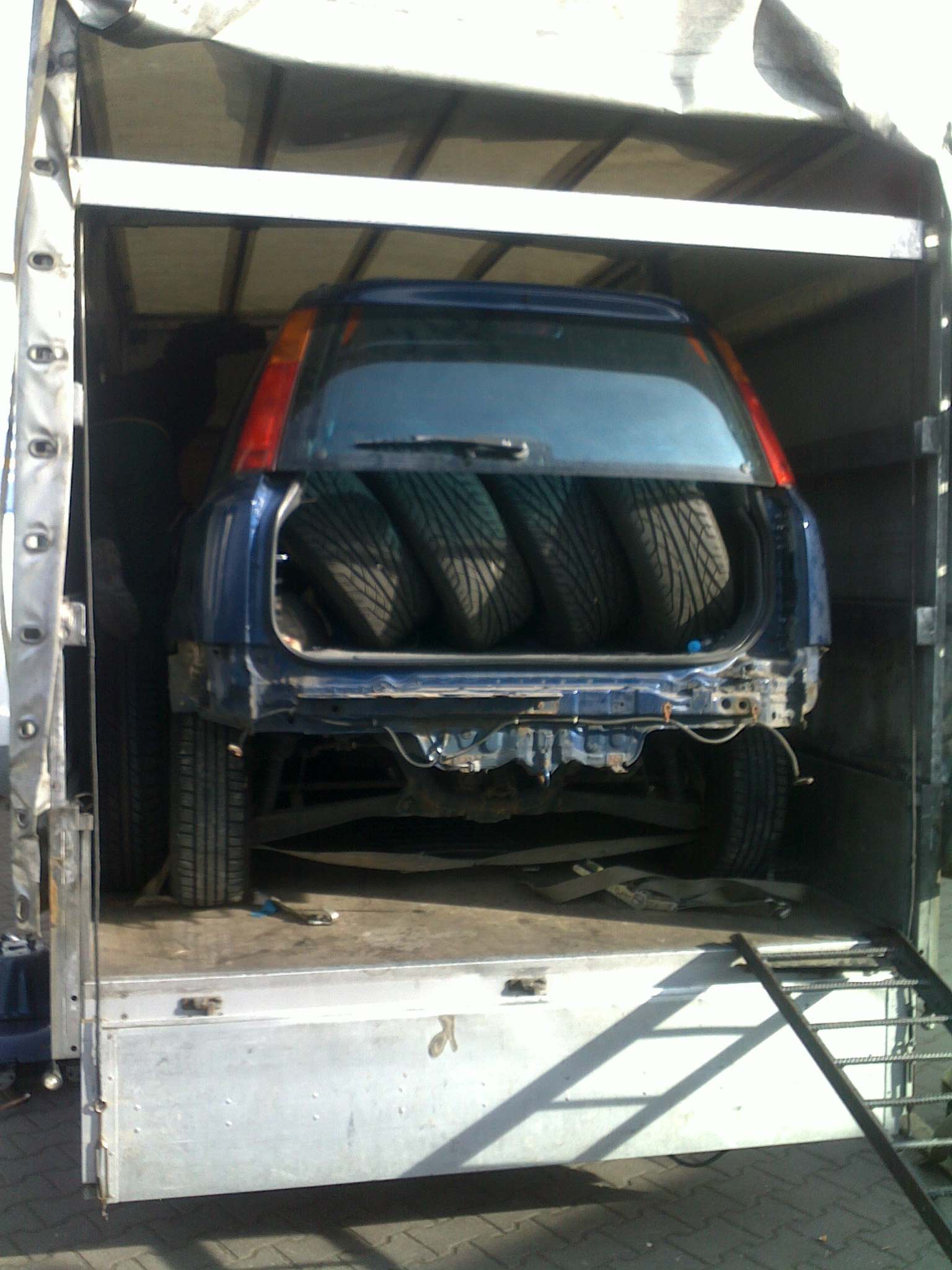 Niebieski samochód osobowy z czterema oponami w bagażniku, zabezpieczony pasami wewnątrz skrzyni ładunkowej samochodu dostawczego z otwartą rampą załadunkową.