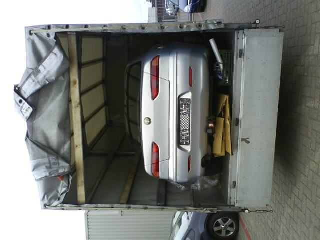 Srebrny samochód osobowy, widoczny z tyłu, zabezpieczony w przestrzeni ładunkowej busa z plandeką; obok kartony i zielony koszyk.